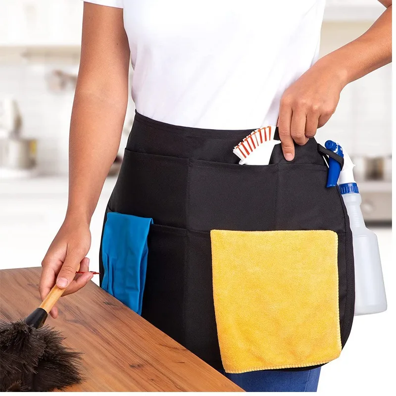 Donna in cucina che indossa il grembiule nero per le pulizie con panno, guanti e spazzola nelle tasche mentre spolvera un tavolo in legno
