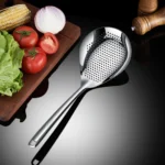 Schiumarola forata in acciaio inox accanto a un tagliere con pomodori, cipolla e macinapepe
