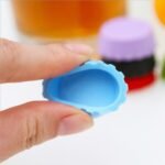 Tappo per bottiglia in silicone blu tenuto tra le dita accanto a un bicchiere di birra e altri tappi